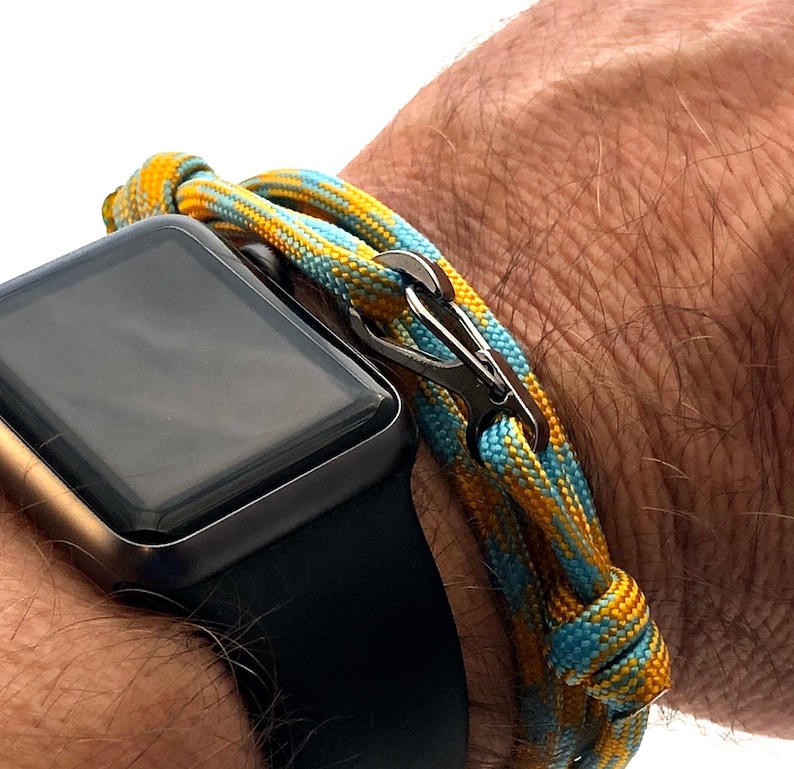 May include: A close-up of a wrist with a black smartwatch and a colorful bracelet. The bracelet is made of woven cord in shades of yellow, blue, and a silver clasp. The watch has a black band and a dark screen.