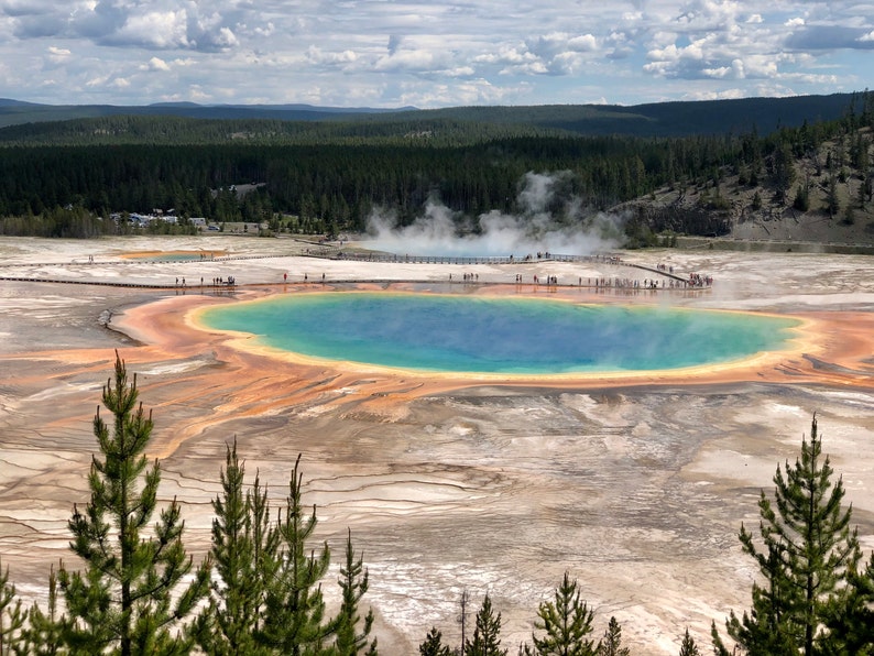 May include: An aerial view of the Grand Prismatic Spring in Yellowstone National Park. The spring features vibrant colors, including blue, orange, and yellow. Steam rises from the water, and a boardwalk allows visitors to walk around the spring.