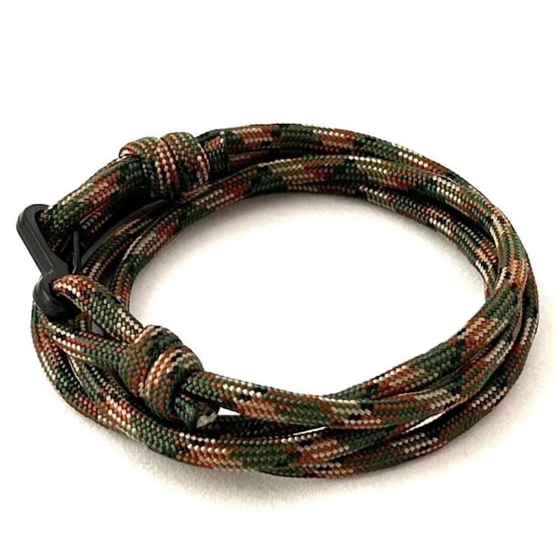May include: Camouflage-patterned paracord bracelet with a black clasp. The bracelet features a mix of green, brown, and white colors, creating a textured appearance. The design is a looped style, suitable for outdoor enthusiasts.