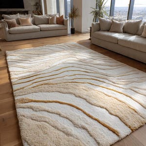 May include: A large, rectangular area rug with a textured, layered design. The rug features a cream-colored base with undulating stripes of beige and gold, creating a flowing, organic pattern. The rug is placed on a hardwood floor in a modern living room.