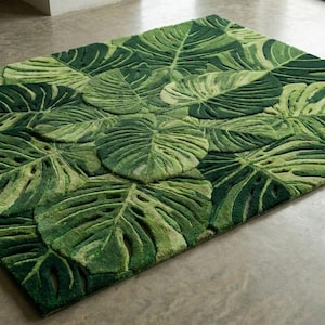 Puede incluir: Una alfombra rectangular verde con un patrón repetido de grandes hojas de monstera estilizadas. Las hojas varían en tonos de verde, creando un efecto texturizado y tridimensional. La alfombra está colocada sobre un suelo gris claro.
