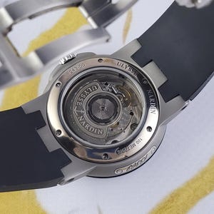 May include: A close-up shot of a high-end wristwatch with a black rubber strap. The watch has a silver-coloured case and a transparent back, revealing the internal mechanisms. The brand name "ULYSSE NARDIN" is displayed.