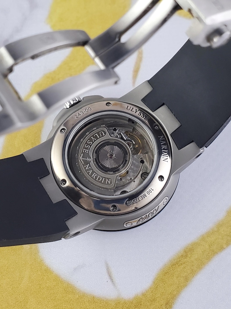 May include: A close-up shot of a high-end wristwatch with a black rubber strap. The watch has a silver-coloured case and a transparent back, revealing the internal mechanisms. The brand name "ULYSSE NARDIN" is displayed.