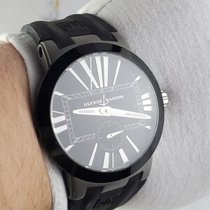 May include: A black Ulysse Nardin watch with a grey face and white Roman numerals. The watch has a black bezel and a black rubber strap. The watch is a luxury item and is designed for men and women.