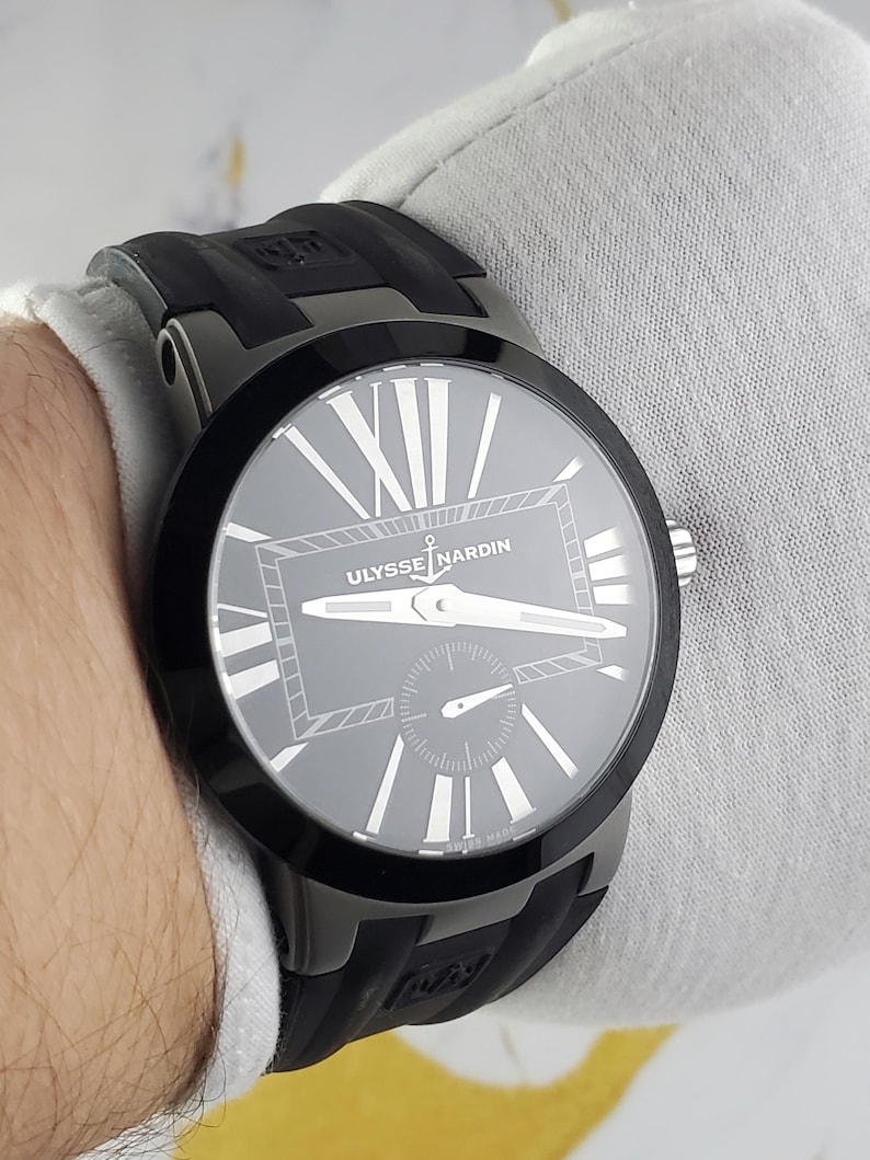 May include: A black Ulysse Nardin watch with a grey face and white Roman numerals. The watch has a black bezel and a black rubber strap. The watch is a luxury item and is designed for men and women.