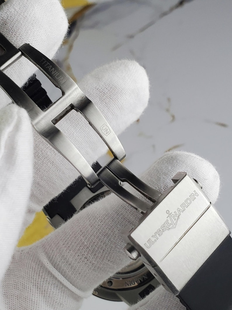 May include: Close-up of a titanium watch clasp. The silver clasp features the word "TITANIUM" and the brand name "ULYSSE NARDIN". The watch band is black. The clasp is being opened, revealing the watch mechanism.