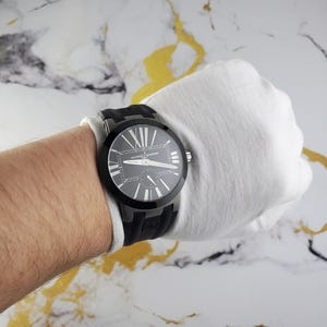 May include: A black Ulysse Nardin watch with a black rubber strap. The watch face is dark with white Roman numerals and hands. The watch is on a wrist covered by a white glove.