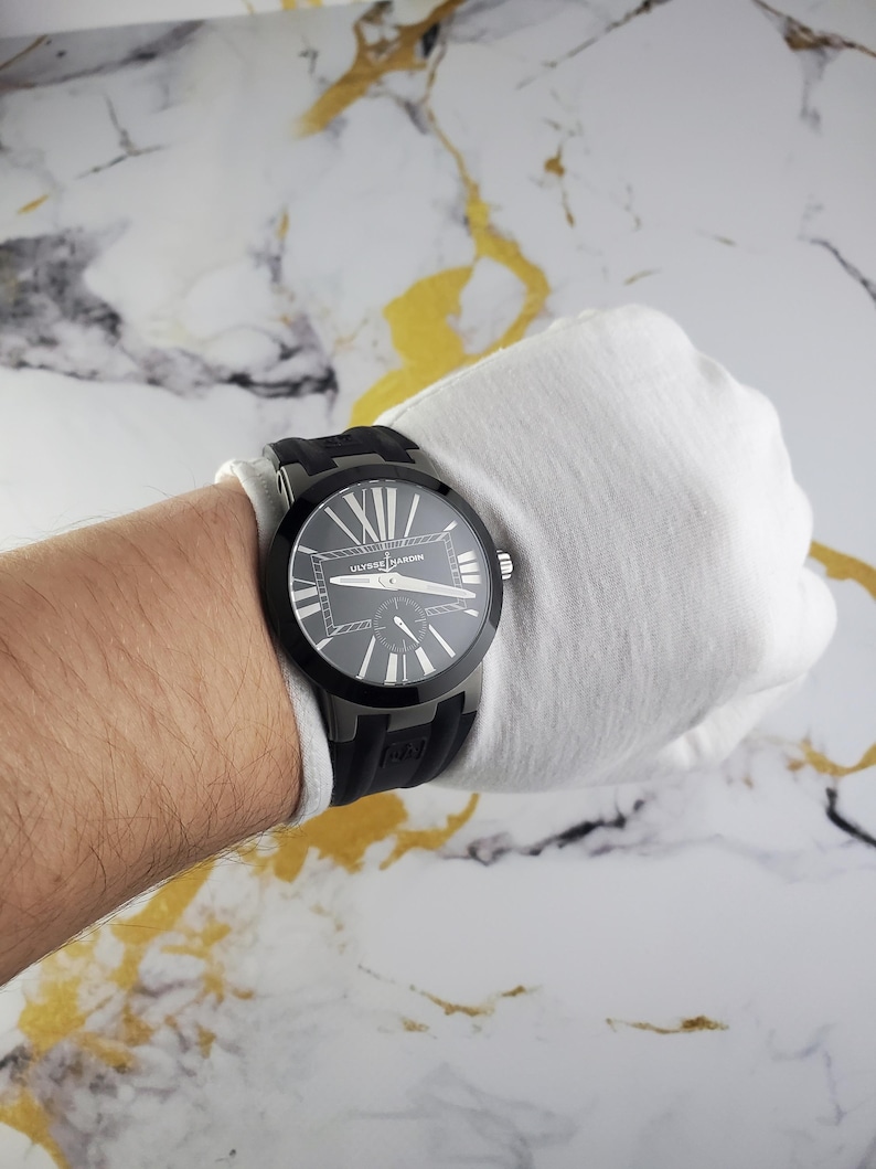 May include: A black Ulysse Nardin watch with a black rubber strap. The watch face is dark with white Roman numerals and hands. The watch is on a wrist covered by a white glove.