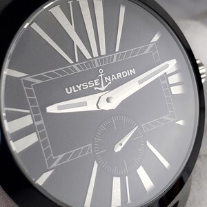 May include: Close-up of a black Ulysse Nardin watch. The grey watch face features silver Roman numerals and hands. The brand name and anchor logo are centered. A small seconds dial is at the bottom. The watch has a black bezel and band.