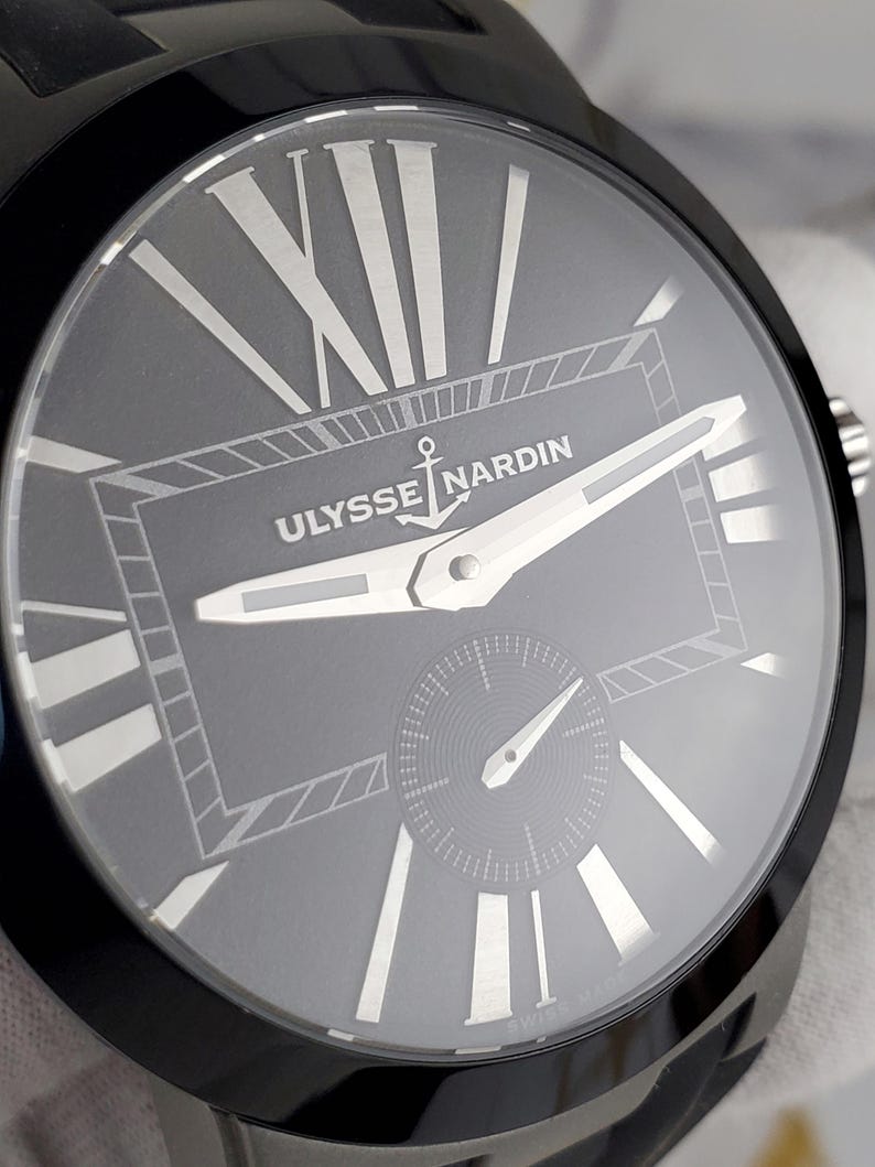 May include: Close-up of a black Ulysse Nardin watch. The grey watch face features silver Roman numerals and hands. The brand name and anchor logo are centered. A small seconds dial is at the bottom. The watch has a black bezel and band.
