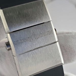 May include: A close-up of a watch clasp with the brand name "ULYSSE NARDIN" engraved on the brushed metal surface. The clasp is rectangular and connects to a black rubber strap.
