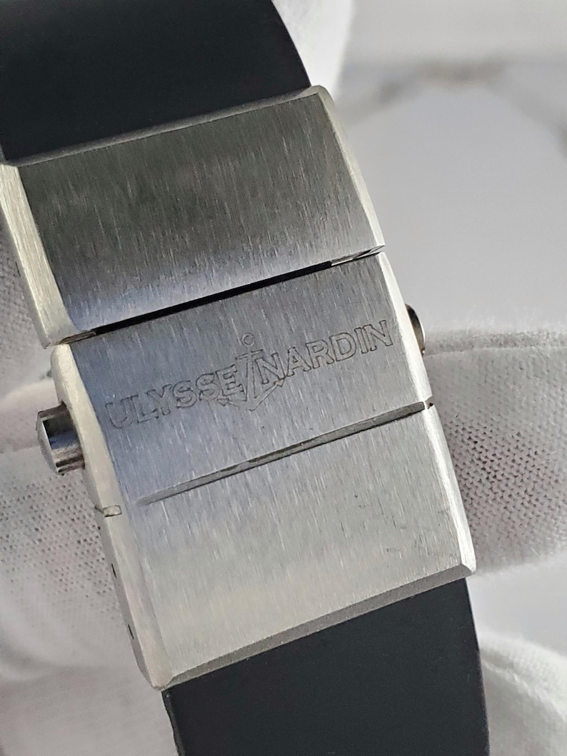 May include: A close-up of a watch clasp with the brand name "ULYSSE NARDIN" engraved on the brushed metal surface. The clasp is rectangular and connects to a black rubber strap.