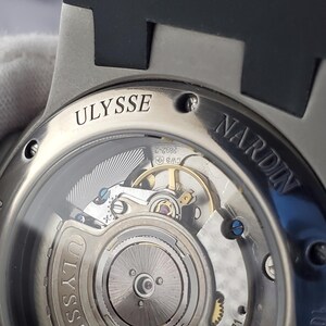 May include: A close-up view of a silver watch with a black strap. The watch's transparent back reveals the complex internal mechanism. The brand names "ULYSSE" and "NARDIN" are engraved on the watch casing.