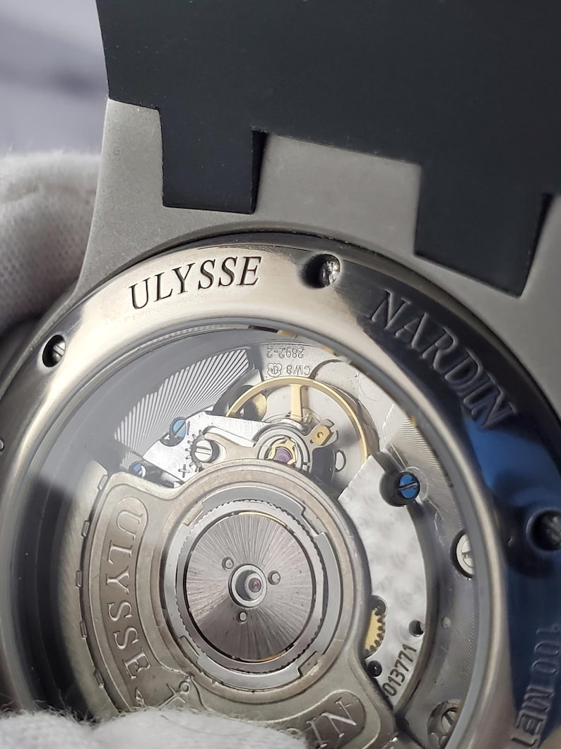 May include: A close-up view of a silver watch with a black strap. The watch's transparent back reveals the complex internal mechanism. The brand names "ULYSSE" and "NARDIN" are engraved on the watch casing.