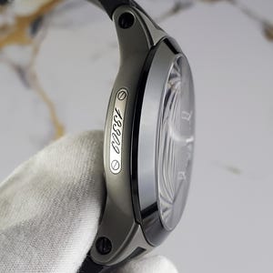 May include: A modern wristwatch with a black strap and a dark grey case. The watch face is visible, and the side of the case displays the number "13309". The watch is held by a gloved hand.