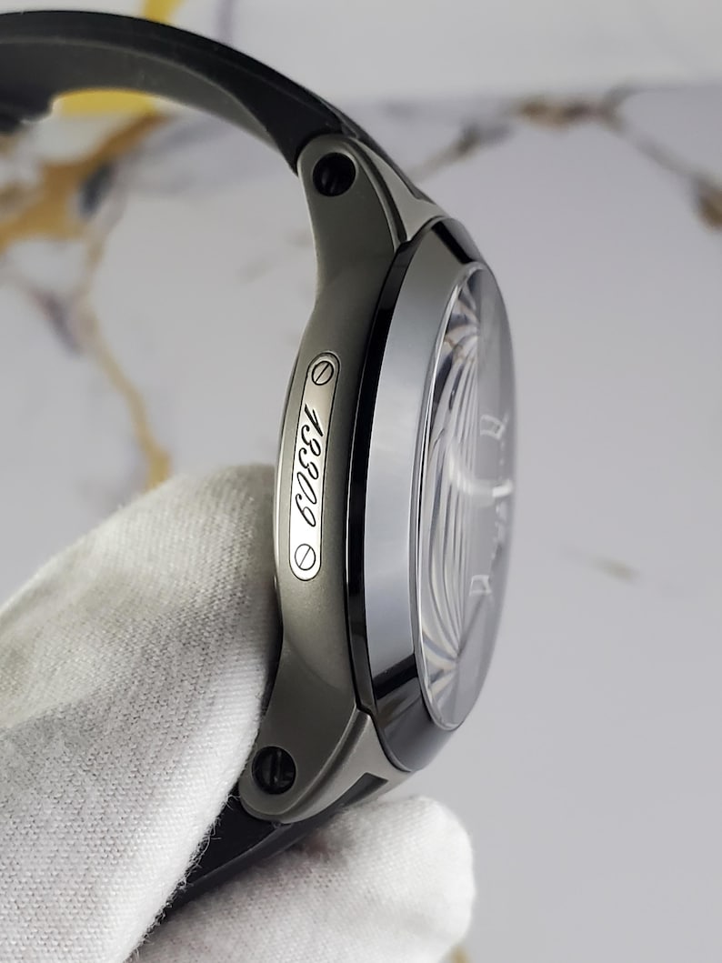 May include: A modern wristwatch with a black strap and a dark grey case. The watch face is visible, and the side of the case displays the number "13309". The watch is held by a gloved hand.