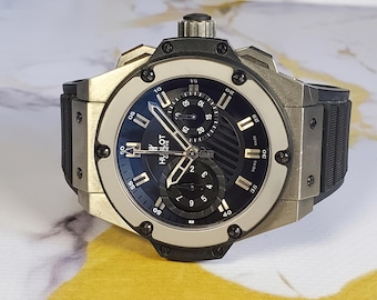 Op de afbeelding: Een luxe polshorloge met een zwarte wijzerplaat en zilverkleurige wijzers en markeringen. Het horloge heeft een zwarte rubberen band en een zilverkleurige kast. De wijzerplaat bevat de merknaam "HUBLOT GENEVE".