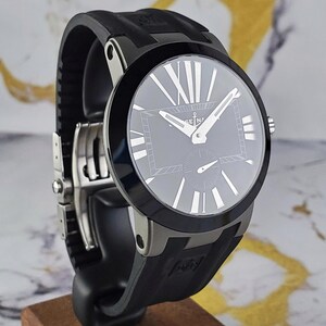May include: A black wristwatch with a round face and a black rubber strap. The watch face has silver hands and Roman numeral hour markers. The watch is displayed on a wooden stand.