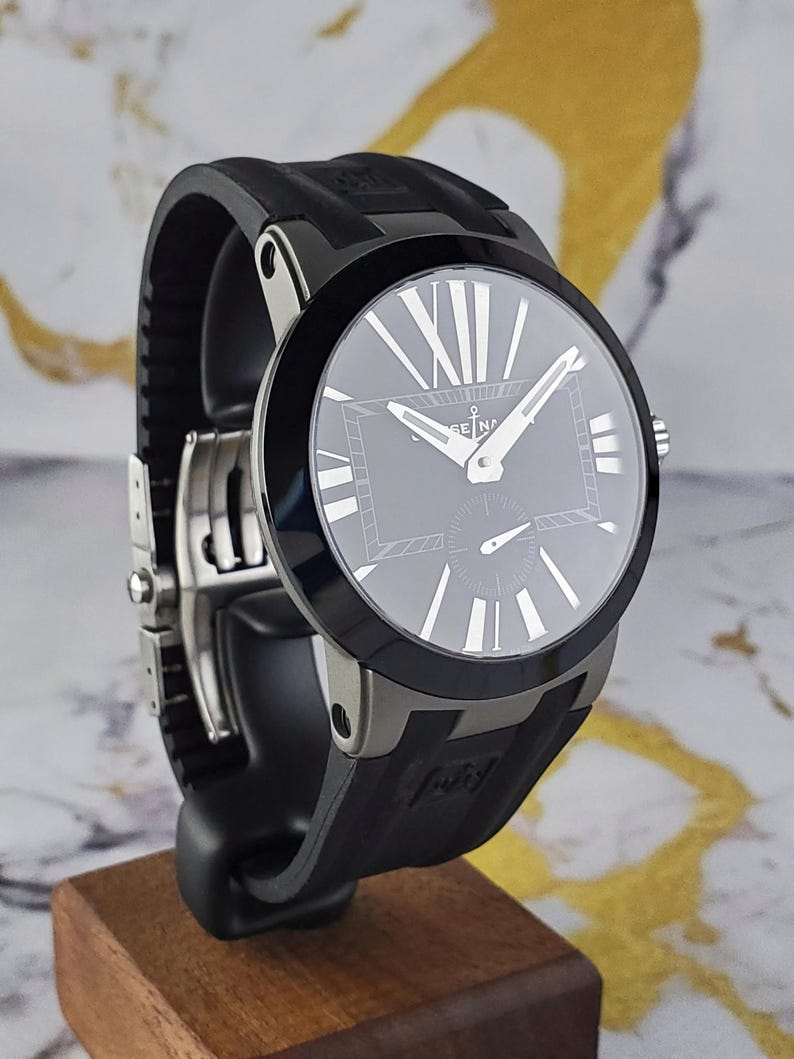 May include: A black wristwatch with a round face and a black rubber strap. The watch face has silver hands and Roman numeral hour markers. The watch is displayed on a wooden stand.