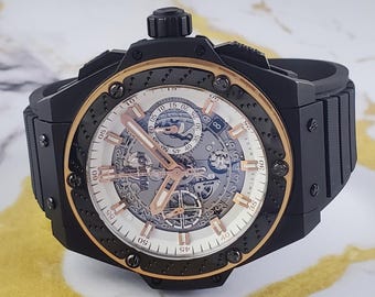 Op de afbeelding: Zwart en goudkleurig luxe polshorloge met een skeleton wijzerplaat. Het horloge heeft een zwarte bezel en band, met goudkleurige accenten rond de wijzerplaat. De wijzerplaat geeft de tijd weer met Romeinse cijfers en markeringen.