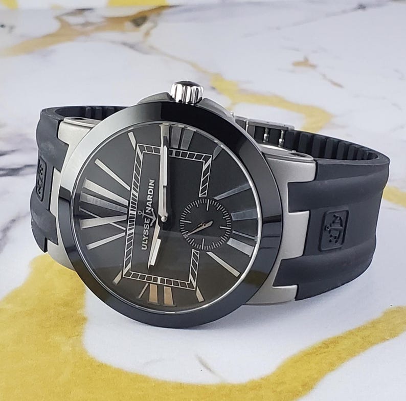 May include: A black Ulysse Nardin watch with a black dial, silver hands, and Roman numeral hour markers. The watch has a black bezel and a black rubber strap. The watch is displayed on a light-coloured surface.