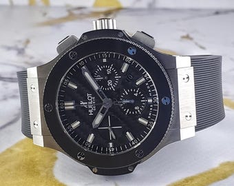 Op de afbeelding: Een zwarte Hublot Geneve horloge met een carbon fiber wijzerplaat en een zwarte rubberen band. Het horloge heeft zilveren accenten, meerdere sub-wijzerplaten en een lunette met minutenmarkeringen. Diameter: ongeveer 4,5 cm.