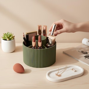 May include: A green, ribbed, rotating makeup organiser filled with lipsticks, makeup brushes, and other beauty products. A hand is reaching in to place a tube of makeup. A small succulent in a white pot and a white tray with jewellery are also on the table.