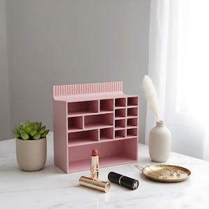 May include: A pink organizer with multiple compartments for cosmetics and accessories. A small succulent in a beige pot, lipsticks, and a gold-toned tray with jewelry are also visible. A white vase with a feather and a black lipstick are also present.