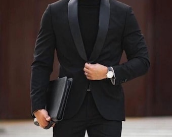 May include: A black suit with a shawl lapel, worn with a black turtleneck and black boots. The suit jacket is buttoned at the waist, and a black leather portfolio is held in the left hand. A silver watch is visible on the left wrist.