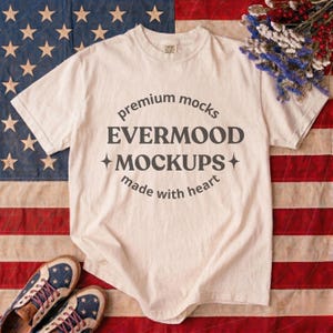 May include: A cream-colored t-shirt with the text "premium mocks EVERMOOD MOCKUPS made with heart" in a circular design. The shirt is laid on an American flag. A pair of blue and white shoes with stars and stripes are at the bottom.
