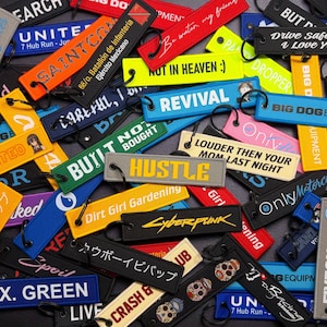 May include: A collection of colorful embroidered keychains with various text designs. Keychains include phrases like "Hustle," "Revival," and "Drive Safe Baby." The keychains are in various colors, including black, yellow, and blue.