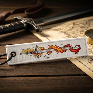 May include: A white rectangular keychain tag featuring a sword engulfed in flames. The sword design is in the center, with orange and red flames extending from both sides. A black key ring is attached to the tag. The background includes a map, compass, and a sword.
