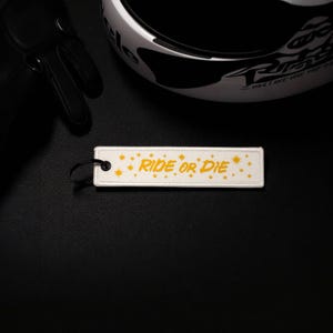 May include: A white rectangular keychain tag with the words "RIDE OR DIE" in yellow, along with yellow starburst designs. The tag is next to a motorcycle helmet and black gloves on a black surface.