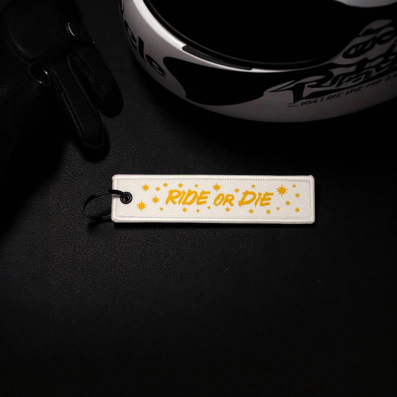 May include: A white rectangular keychain tag with the words "RIDE OR DIE" in yellow, along with yellow starburst designs. The tag is next to a motorcycle helmet and black gloves on a black surface.