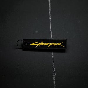 May include: Black rectangular keychain with the word "Cyberpunk" in yellow stylized lettering. The keychain has a black metal ring attached to one end. The background is a dark, textured surface.