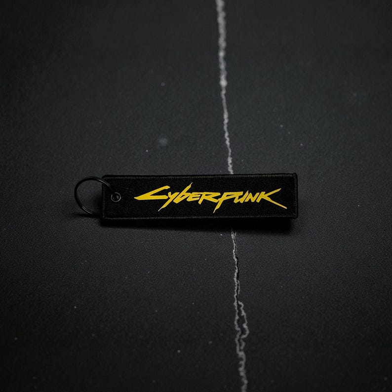 May include: Black rectangular keychain with the word "Cyberpunk" in yellow stylized lettering. The keychain has a black metal ring attached to one end. The background is a dark, textured surface.