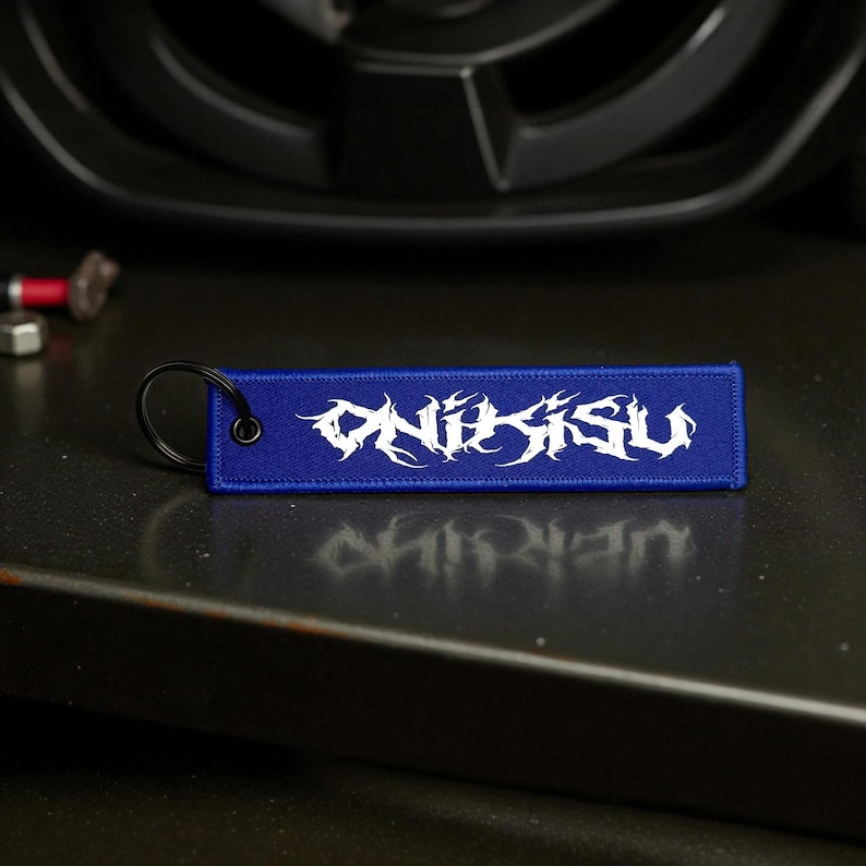 May include: A blue rectangular keychain tag with the word "ONIKISU" in white, stylized lettering. The tag has a black metal ring for attachment. The background includes a car wheel and other automotive parts.