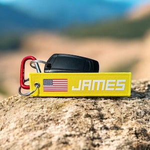 May include: A yellow keychain with the name "JAMES" in white block letters and an American flag graphic. The keychain is attached to a red carabiner clip and a car key. The background is blurred.