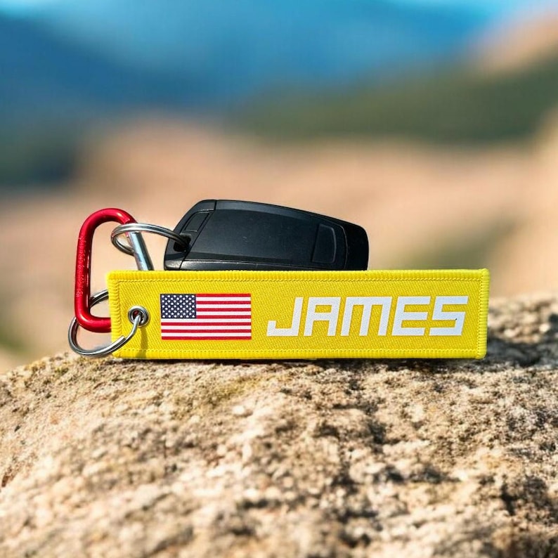 May include: A yellow keychain with the name "JAMES" in white block letters and an American flag graphic. The keychain is attached to a red carabiner clip and a car key. The background is blurred.