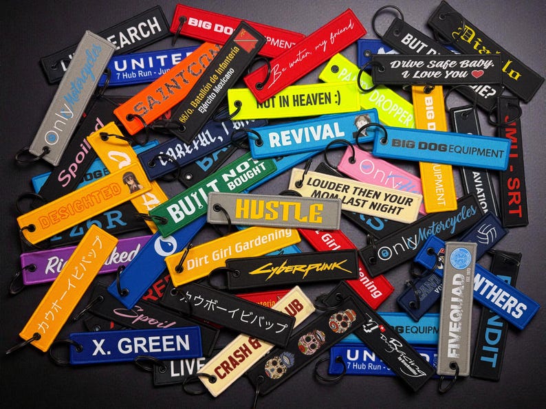 May include: A collection of colorful embroidered keychains with various text designs. Keychains include phrases like "Hustle," "Revival," and "Drive Safe Baby." The keychains are in various colors, including black, yellow, and blue.