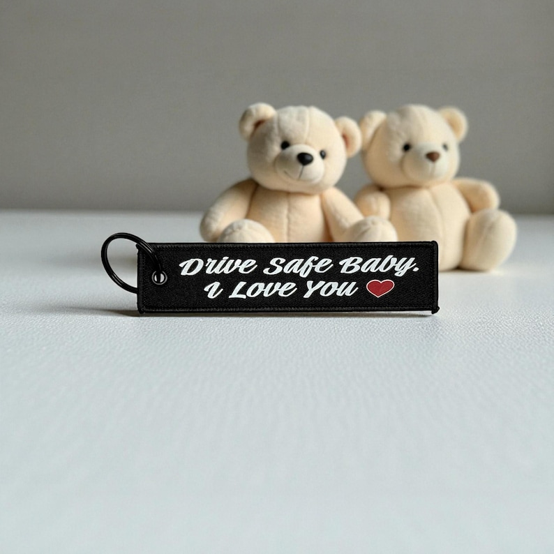 May include: A black keychain with the text "Drive Safe Baby. I Love You" in white, accompanied by a red heart. Two beige teddy bears sit in the background. The keychain has a metal ring.