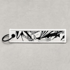 May include: A rectangular white keychain tag with a black illustration of a character's eyes. The tag has a black metal ring attached to one end. The illustration features detailed lines and shading, creating a dramatic effect. The tag is likely made of fabric.