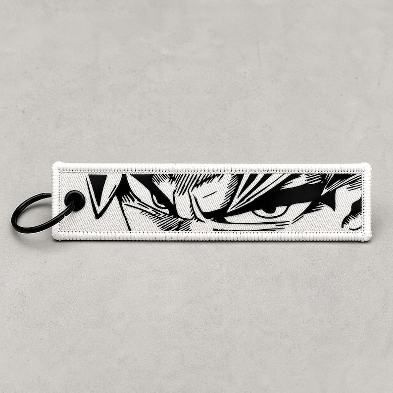 May include: A rectangular white keychain tag with a black illustration of a character's eyes. The tag has a black metal ring attached to one end. The illustration features detailed lines and shading, creating a dramatic effect. The tag is likely made of fabric.