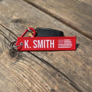 May include: Red rectangular keychain with the text "K. SMITH" in white, along with a small American flag graphic. The keychain is attached to a silver keyring and a red carabiner clip. A black car key is visible in the background.