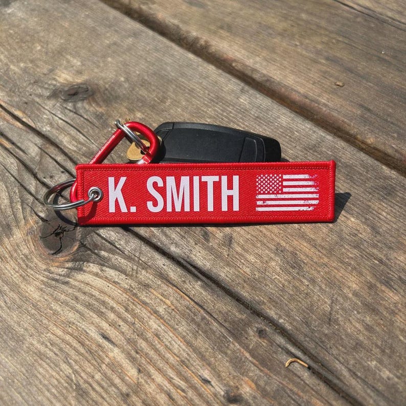 May include: Red rectangular keychain with the text "K. SMITH" in white, along with a small American flag graphic. The keychain is attached to a silver keyring and a red carabiner clip. A black car key is visible in the background.