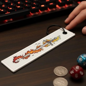 May include: A white rectangular keychain featuring a sword design engulfed in flames. The keychain has a black ring and is placed on a dark wooden surface, next to dice, coins, and a keyboard.