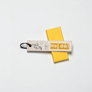 May include: A rectangular keychain with the text "Hej d&aring;!" in yellow, featuring a black and white illustration of a car accident and a person. The keychain has a black ring and is placed on a yellow box.