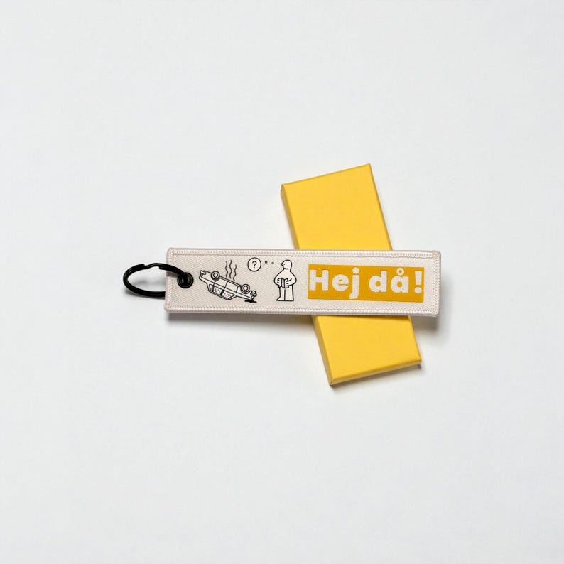 May include: A rectangular keychain with the text "Hej d&aring;!" in yellow, featuring a black and white illustration of a car accident and a person. The keychain has a black ring and is placed on a yellow box.