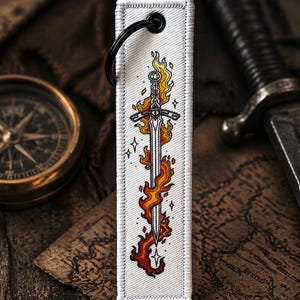 May include: A white rectangular keychain with a black ring features a sword engulfed in flames. The sword design has a detailed hilt and blade, with orange and yellow flames. The keychain is on a wooden surface with a compass and a sword in the background.