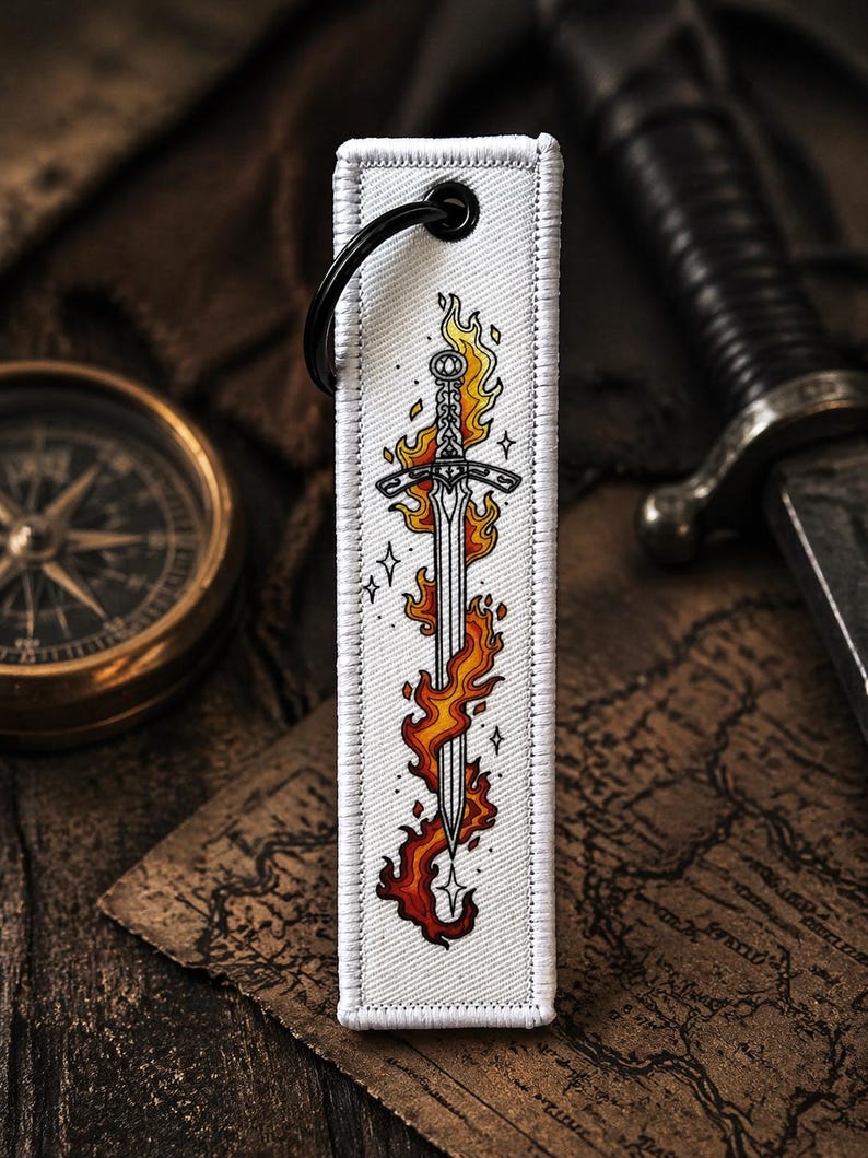 May include: A white rectangular keychain with a black ring features a sword engulfed in flames. The sword design has a detailed hilt and blade, with orange and yellow flames. The keychain is on a wooden surface with a compass and a sword in the background.